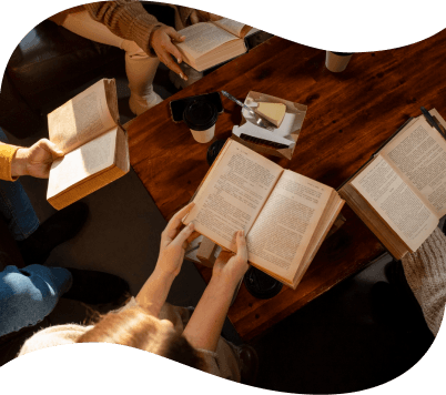 A group of people sitting around a wooden table, each holding an open book, with coffee cups and pastries nearby.