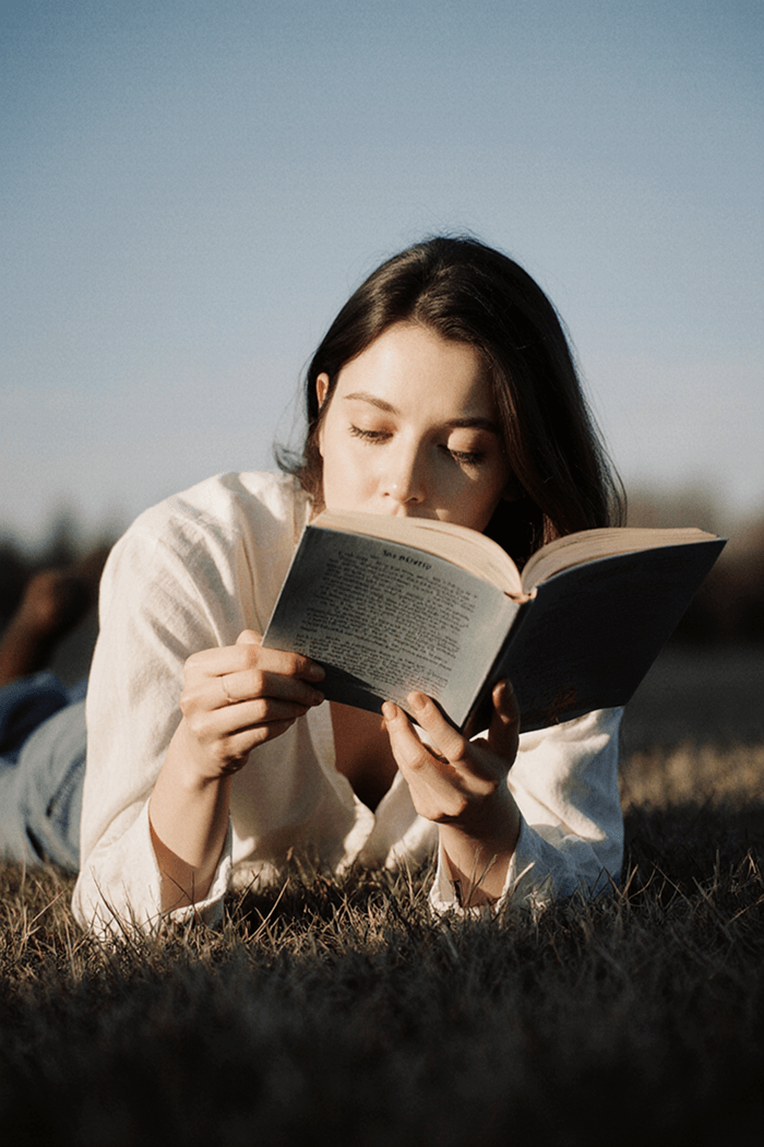 A young woman lying in the grass on a sunny day, deeply focused while reading a book.